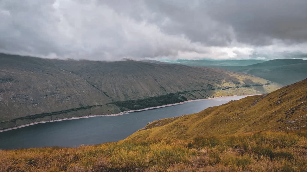 Panoramisch uitzicht op de Schotse Hooglanden, weelderig groene valleien en een vredig meer onder een bewolkte hemel.