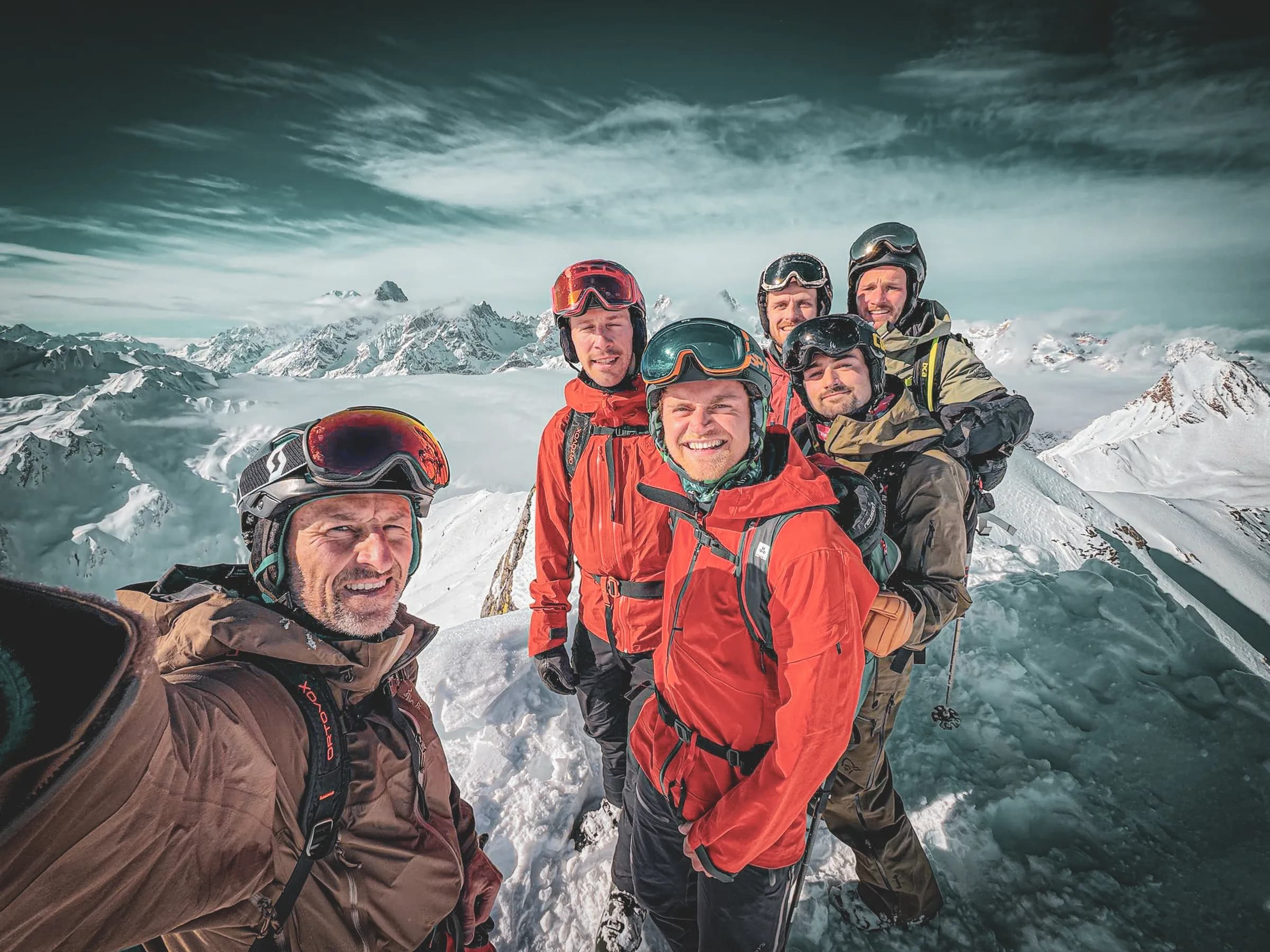A group of five climbers posing for a selfie at the top of a snow-covered mountain. They are wearing colourful ski clothing, helmets and goggles. In the background, majestic peaks tower above them.