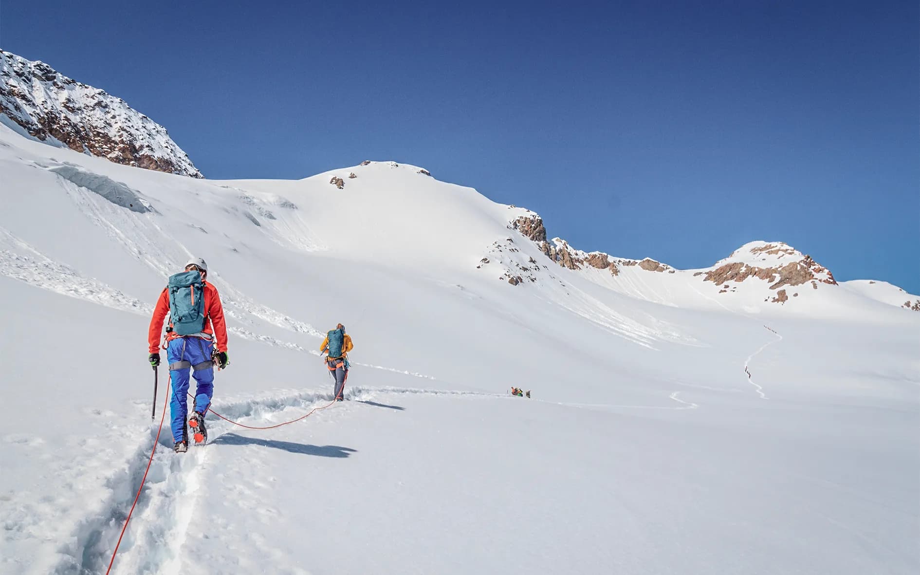 A group of climbers advance over vast snow-covered terrain, pulling safety ropes. They are equipped with cold-weather clothing, rucksacks and ice axes. In the background can be seen
