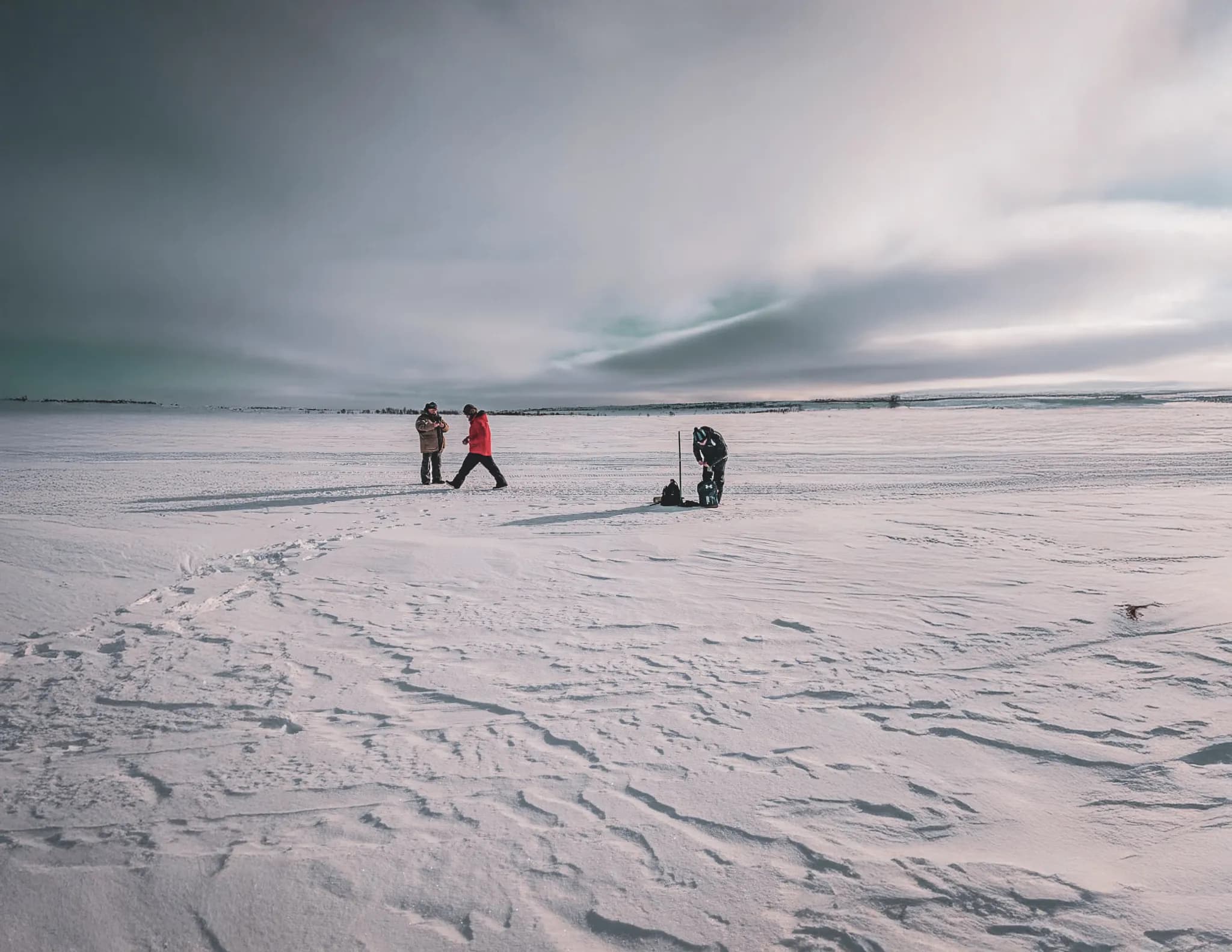 A snow-covered landscape stretches as far as the eye can see, with two people in conversation on the left, one in light clothing and the other in red. On the right, a third person is working with a piece of equipment.