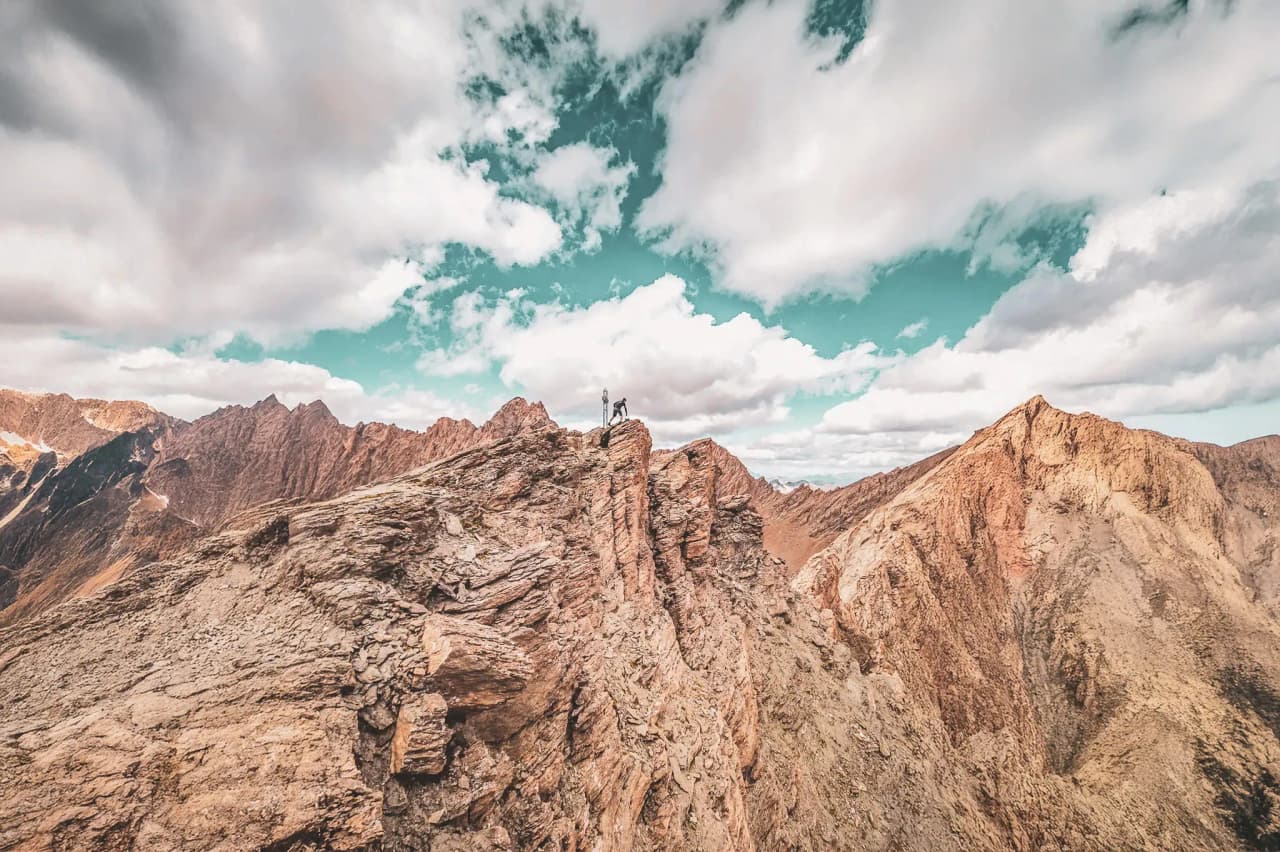 A hiker and his dog at the top of a mountain ridge, surrounded by Alpine scenery.