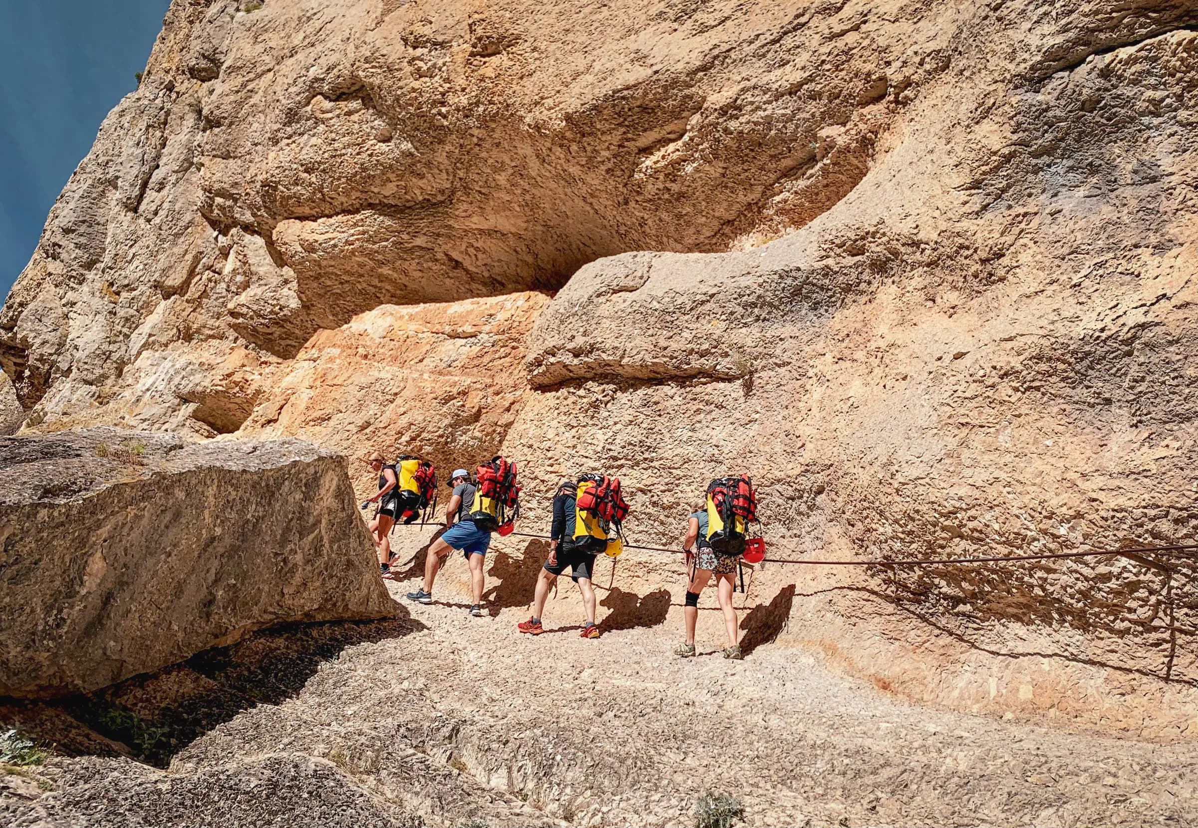 A group of hikers follow a rocky path, framed by an imposing cliff. Each participant carries a large rucksack and is equipped for climbing, with clothing suited to the conditions.