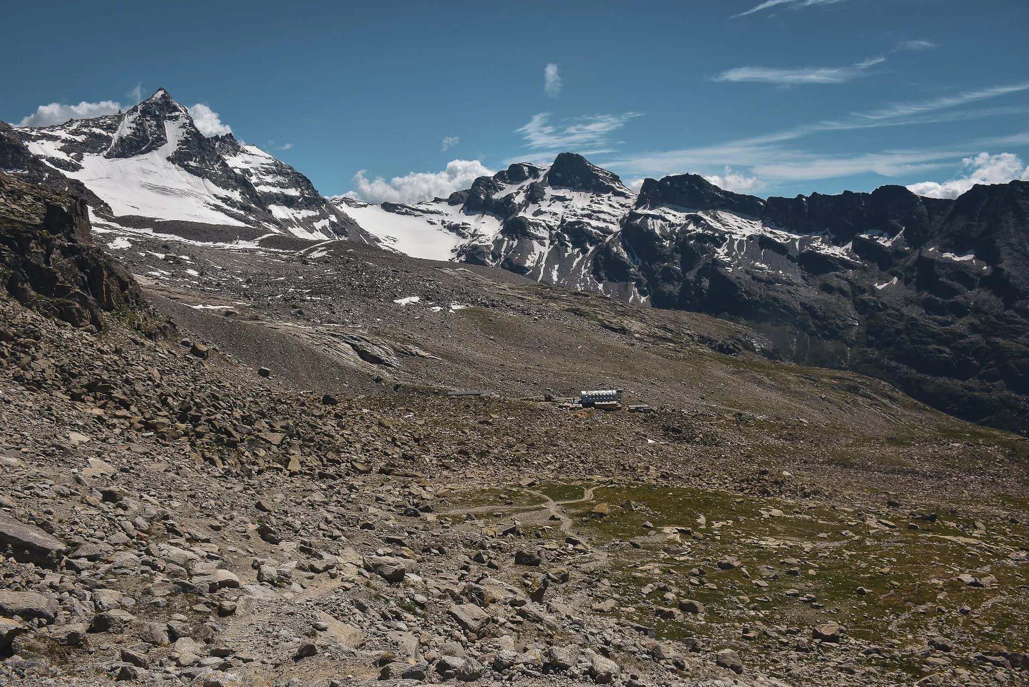 Een berglandschap met besneeuwde toppen onder een helderblauwe lucht. Op de voorgrond een ruig, rotsachtig terrein met verspreide stenen en gras. In de verte staat een berghut.