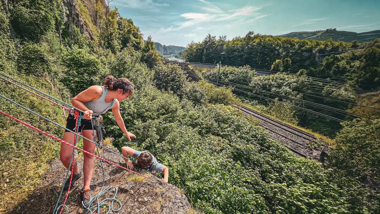 An instructor guides a beginner climber on a green cliff in Yvoir, Belgium.