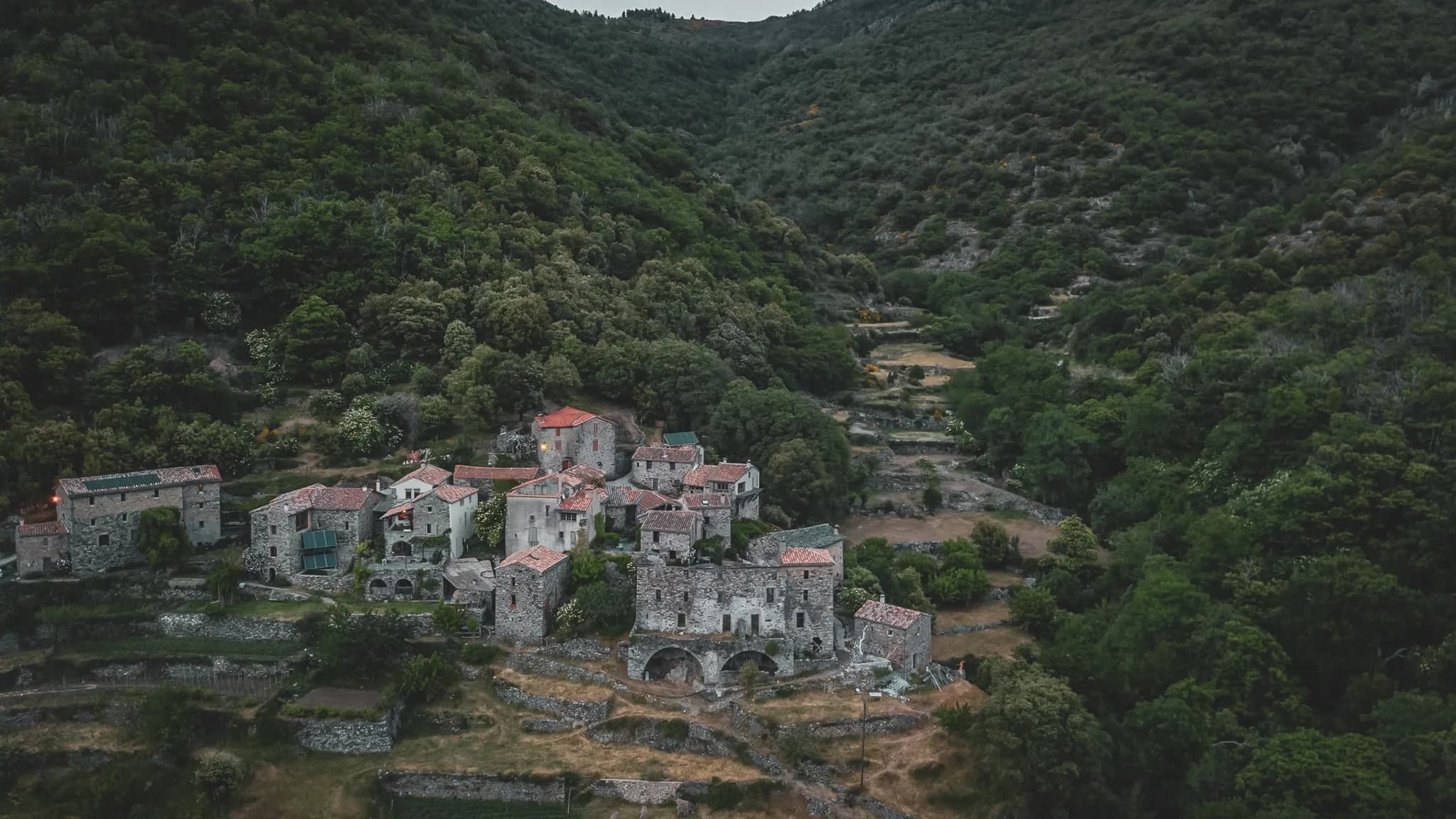 A picturesque village nestling in a green valley, surrounded by hills covered in lush forests. The houses, built of stone with red-tiled roofs, are scattered around the village.
