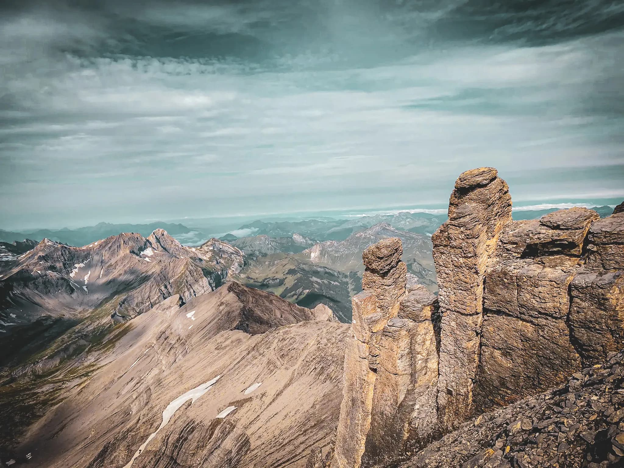 An impressive mountain landscape with steep rock formations in the foreground. The mountains in the background are of varying shades of grey and brown, overgrown with vegetation and dotted with snow. Visit