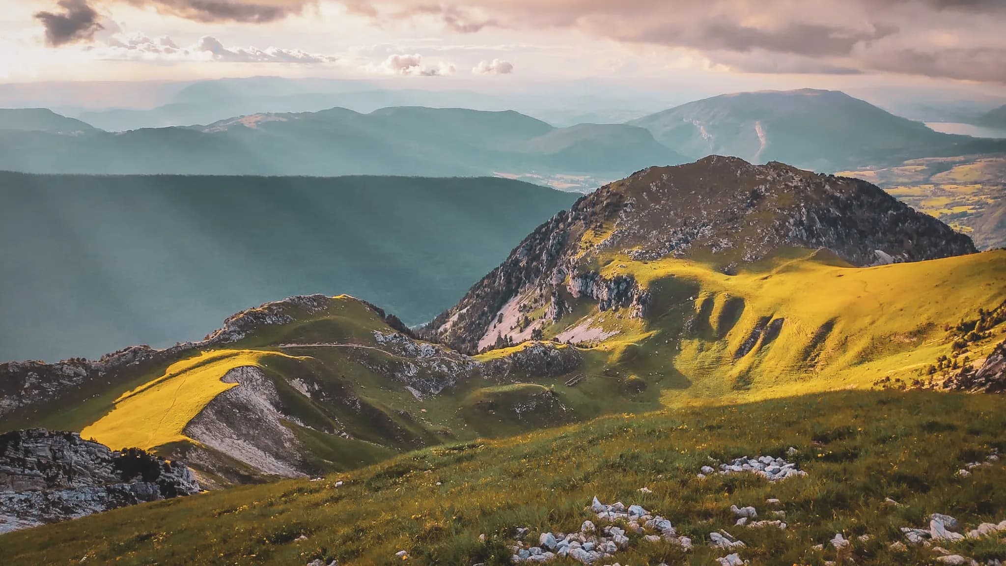 Un paysage montagneux avec des collines verdoyantes et des formations rocheuses. La lumière du soleil perce à travers des nuages, créant des jeux d'ombre et de lumière sur le terrain. En arrière-plan, on aperçoit des