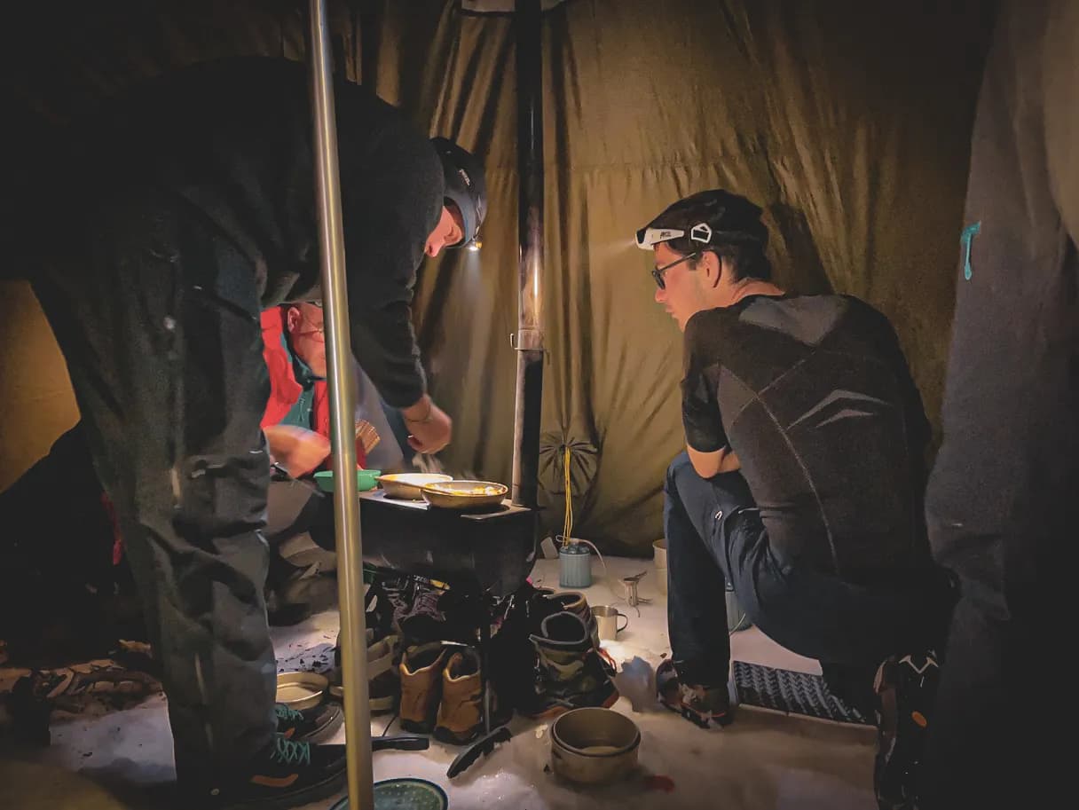 A dark interior, probably a tent, where two people are fussing around a small stove. One is leaning towards the stove, while the other is seated, carrying a headlamp. From