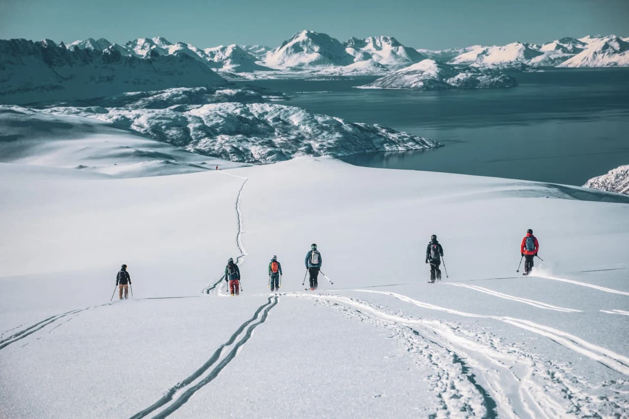 A snowy landscape with a group of six climbers walking on a slope. The immaculate snow is criss-crossed by tracks left by their skis, leading to an impressive mountainous horizon. In the background