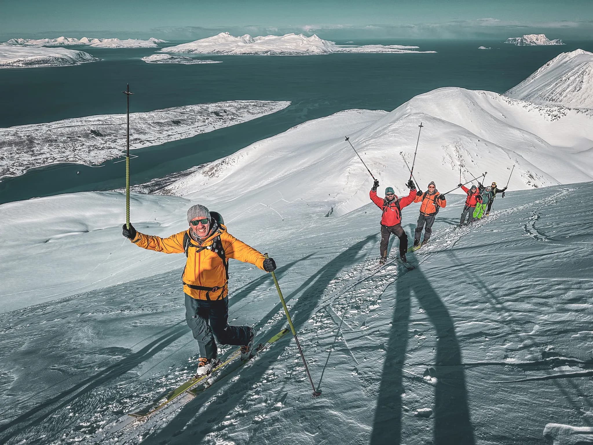 Un groupe de skieurs en montagne, progressant sur une pente enneigée. Au premier plan, un homme souriant en veste orange tient un bâton de ski au-dessus de sa tête, tandis que les autres skieurs, vêtus de
