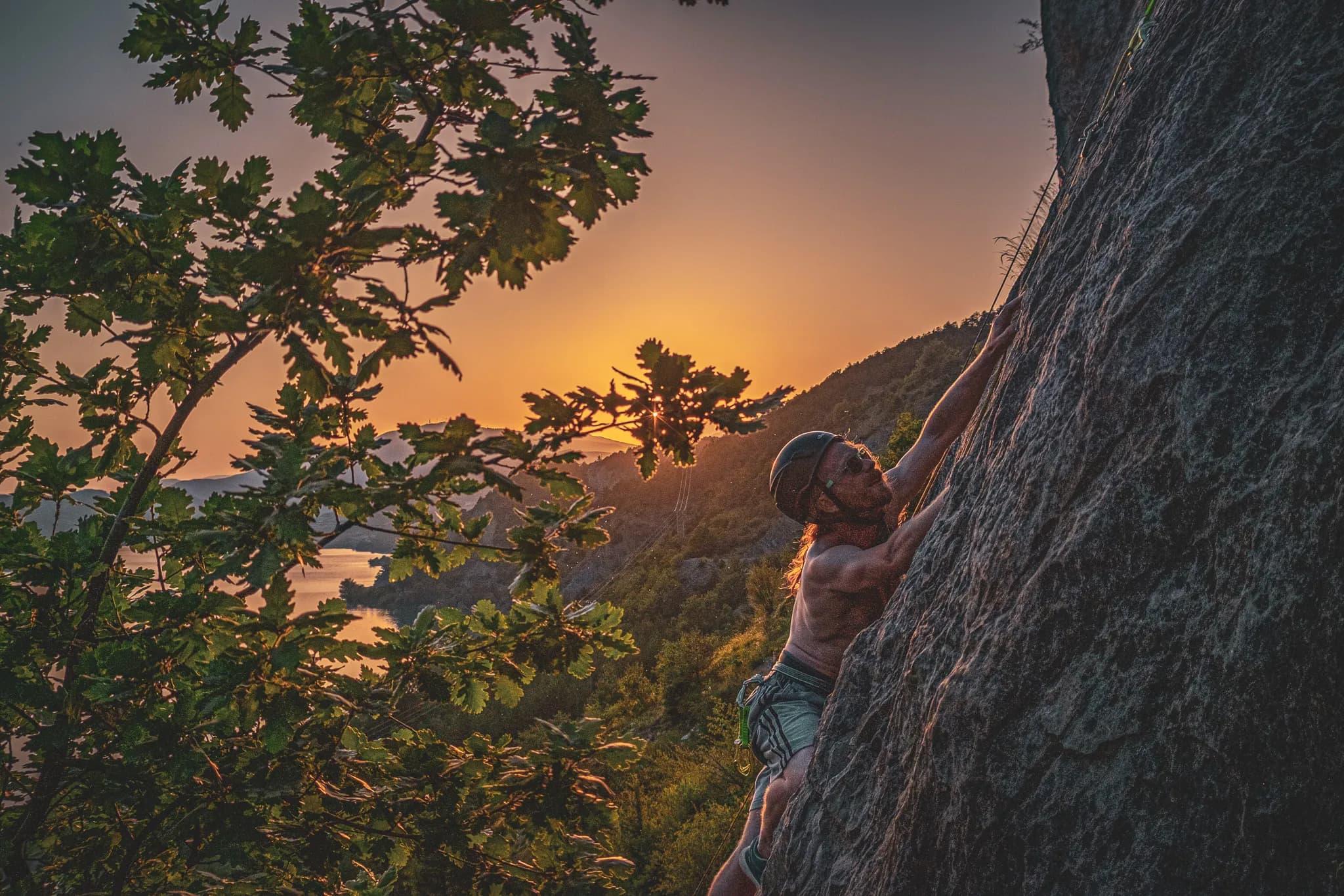 A climber climbs a rock face against the light, while the sun sets on the horizon, creating a warm, golden atmosphere. Green leaves frame the scene, adding to the warmth of the atmosphere.