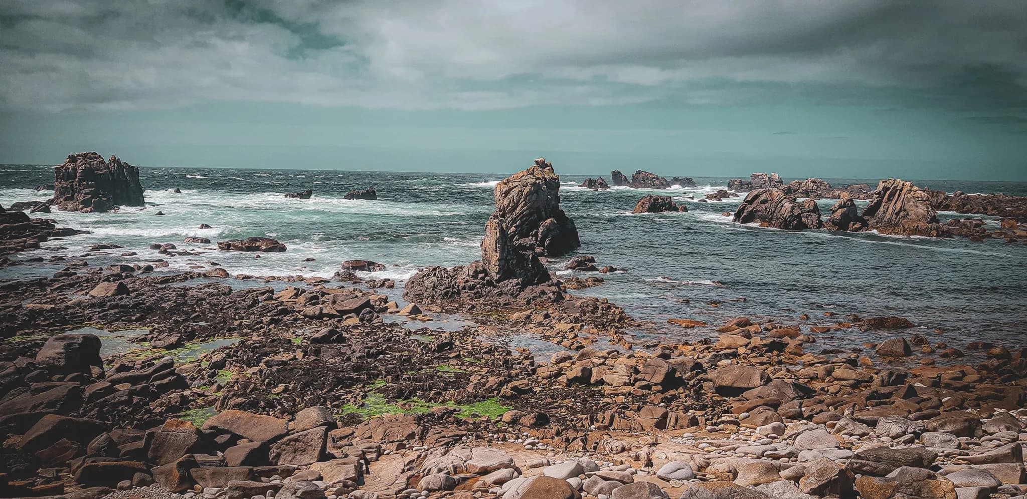 A wild coastal landscape with rocks rising out of the sea. Waves crash against the rock formations, creating a contrast with the blue-green water. The shoreline is dotted with pebbles and