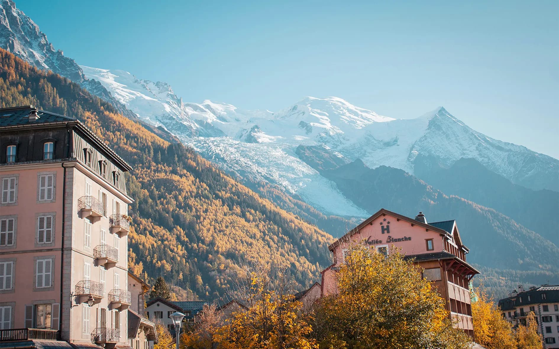 A picturesque alpine landscape, with majestic snow-capped mountains in the background. In the foreground, chalet-style buildings, one of which features a pink façade with traditional architectural elements.
