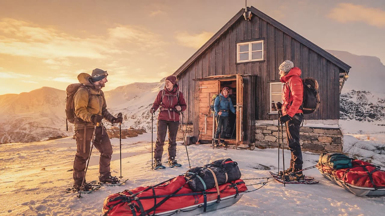Een groep sneeuwschoenwandelaars voor een chalet in Lapland, onder een goudkleurige hemel.
