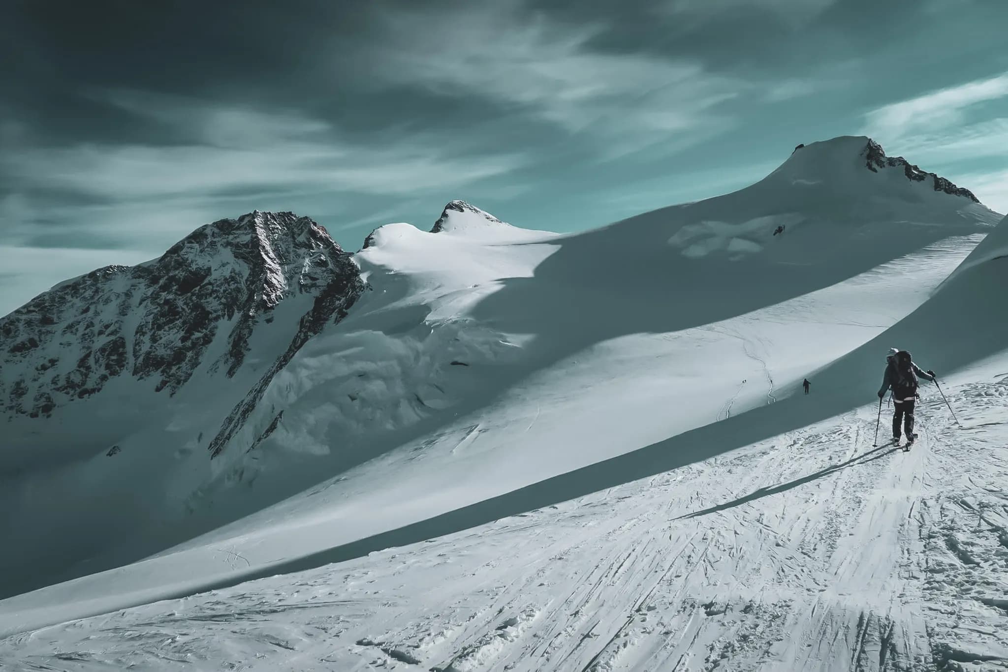 Een besneeuwd berglandschap met majestueuze bergtoppen onder een bewolkte hemel. Een skiër waagt zich op een helling en baant zich een weg door de verse sneeuw. Op de achtergrond zijn andere wandelaars te zien,