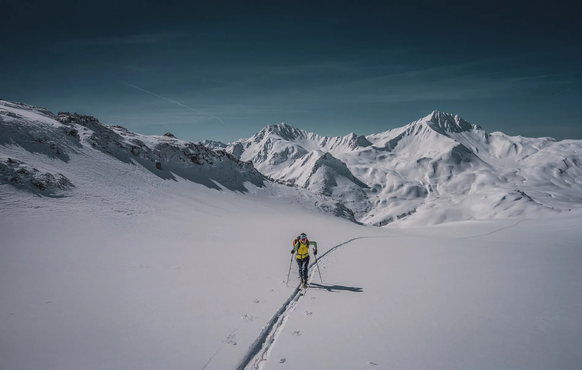 A lone skier crosses a vast snow-covered landscape, leaving tracks in the immaculate snow behind him. Majestic mountains rise up in the distance, their peaks covered in snow. The sky is blue