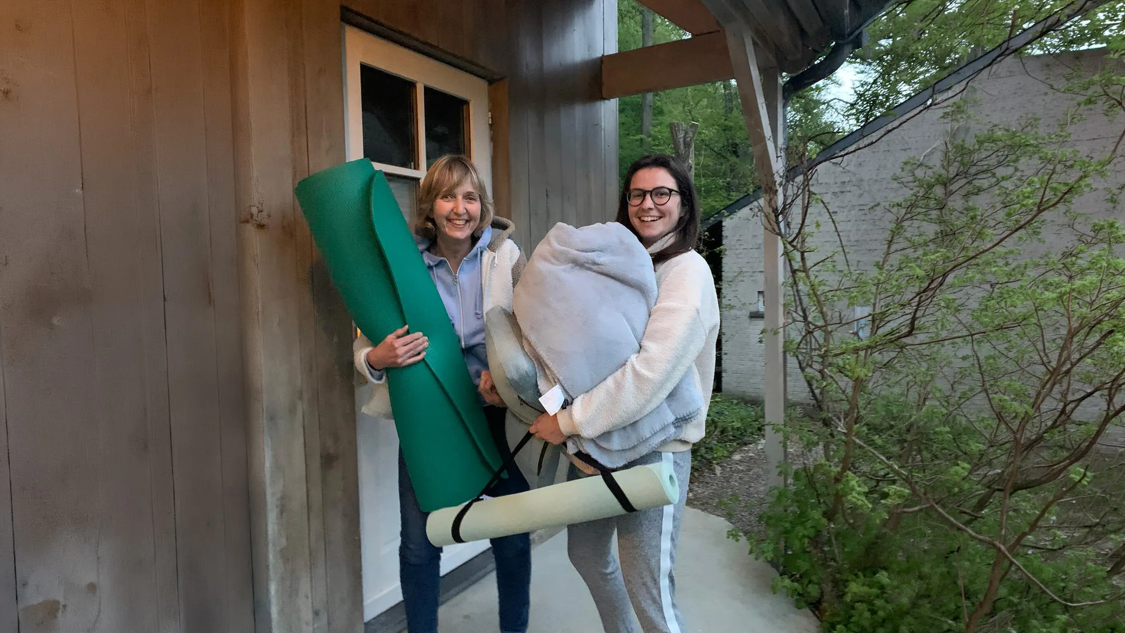 Deux femmes souriantes portant des tapis de yoga et des couvertures, prêtes pour une retraite en nature.