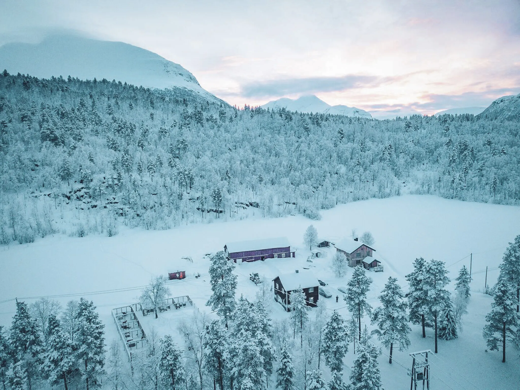 Un paysage hivernal couvert de neige, avec une vaste forêt de conifères bordant un lac gelé. Des structures en bois, dont plusieurs maisons et un bâtiment de ferme, sont éparpillées dans le paysage. Les arbres sont rec
