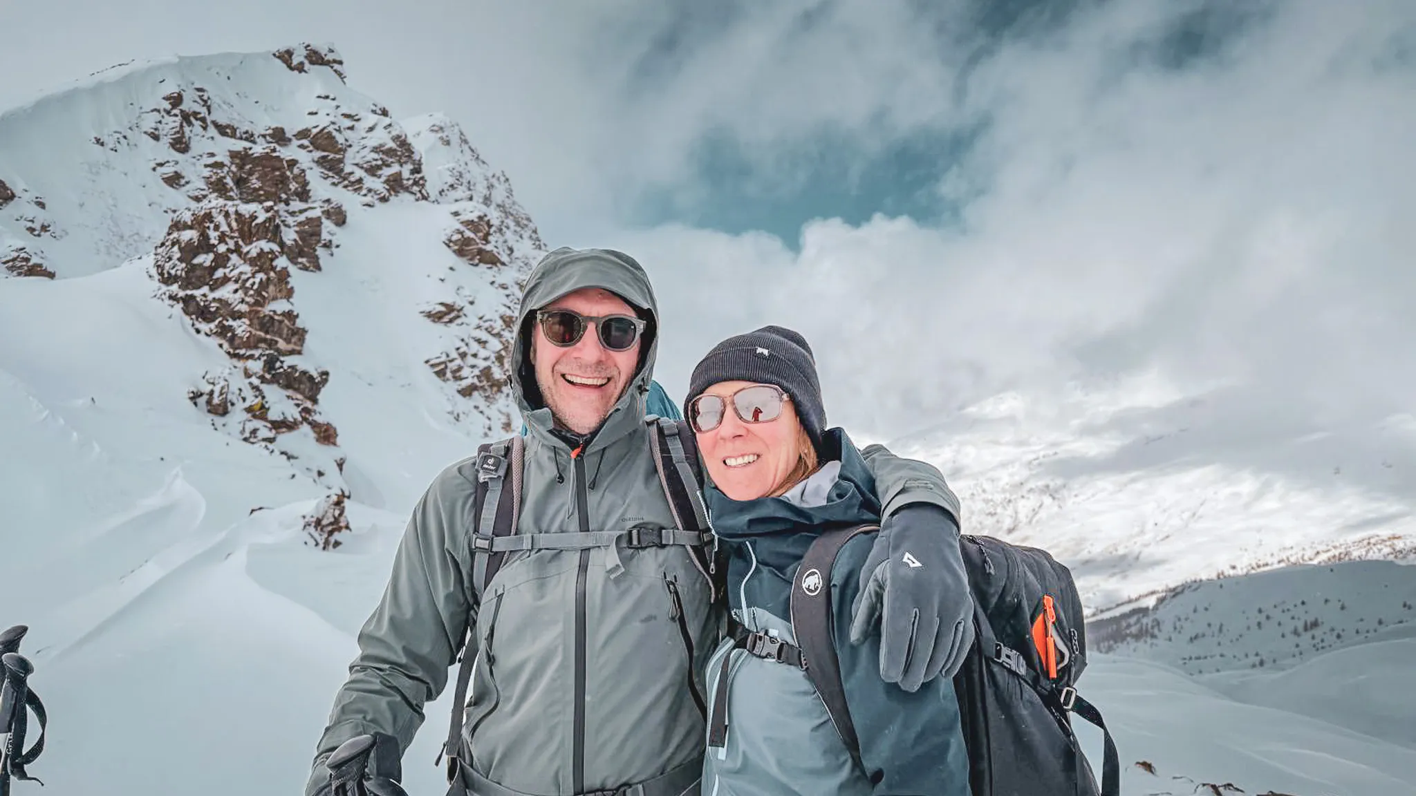 Un couple souriant en ski de randonnée, avec des sommets enneigés en arrière-plan.