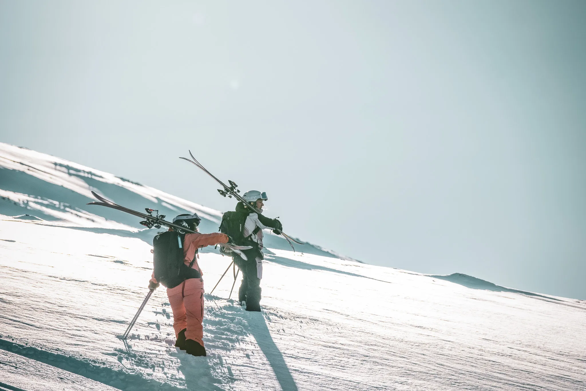 Un couple de skieurs se tient sur une pente enneigée, avec des skis sur l'épaule et des sacs à dos. L'un porte une combinaison orange, tandis que l'autre est vêtu de vêtements en noir et blanc. L'ar