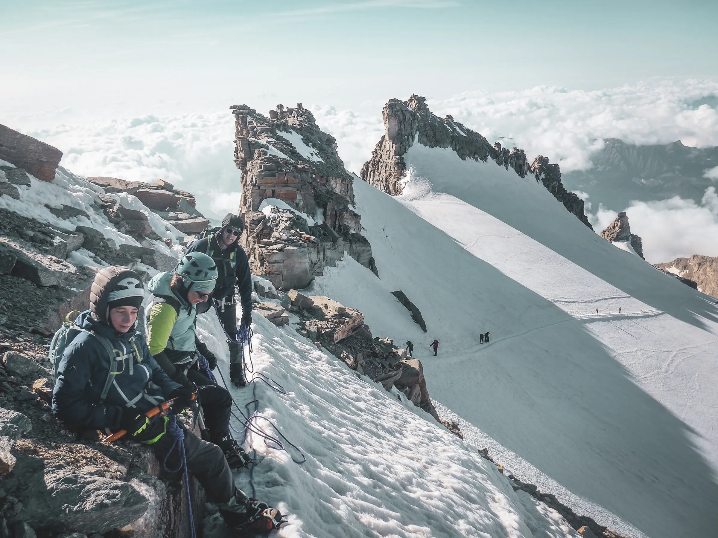 Mountaineers on a snow-covered summit, with wispy clouds and majestic peaks in the background.