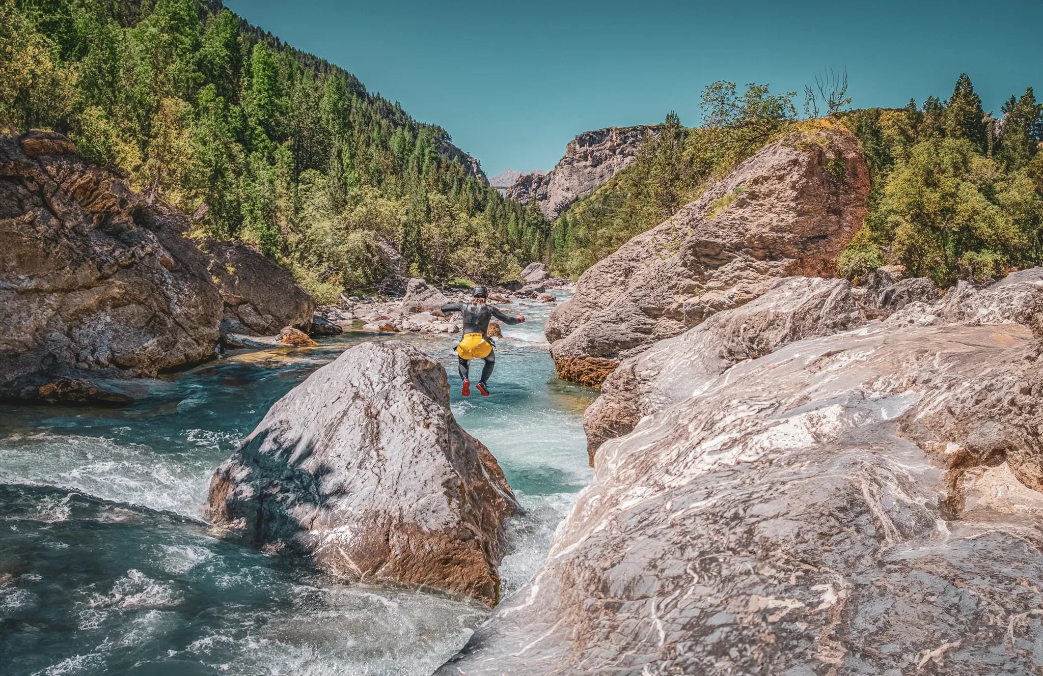 An adventurer in a wetsuit jumps between rocks on the banks of an alpine river, surrounded by lush greenery.