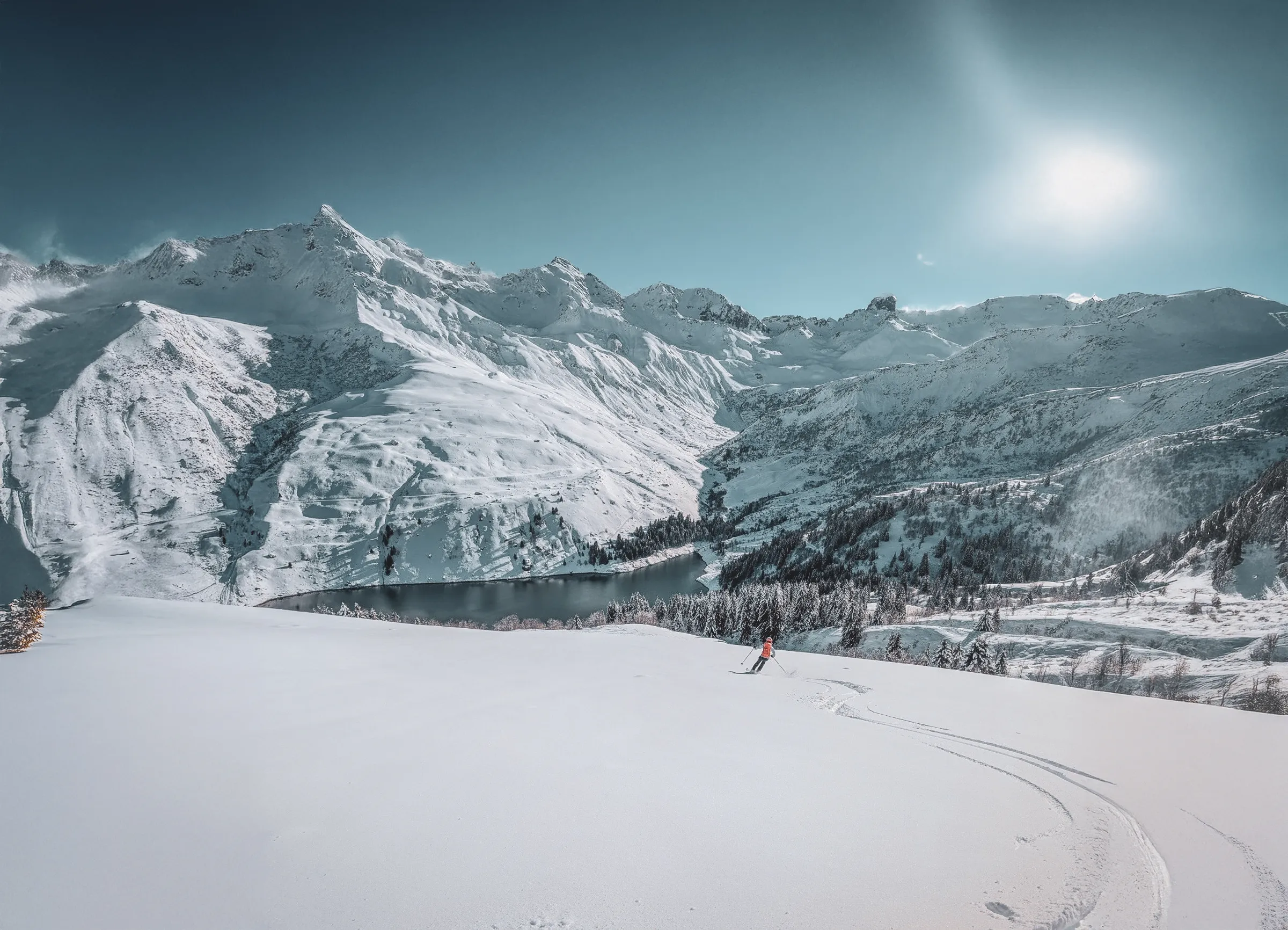 An Alpine landscape in winter, with snow-capped mountains towering majestically beneath a clear blue sky. In the foreground, an expanse of untouched snow bears witness to recent snowfalls. A sk