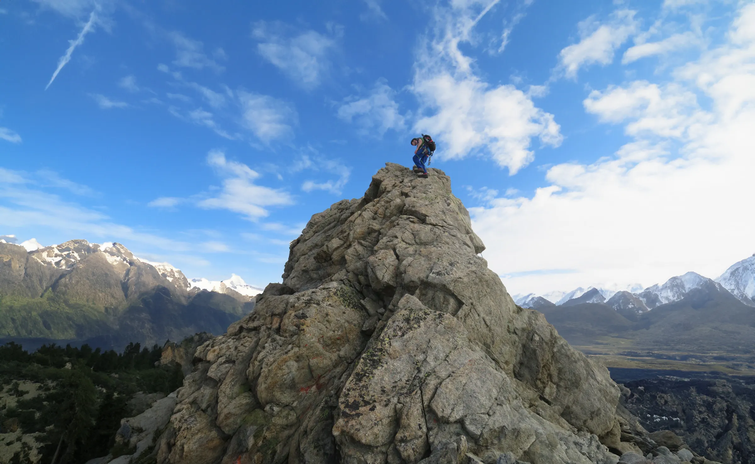 A climber on a majestic crag, facing snow-capped mountains and a brilliant blue sky.