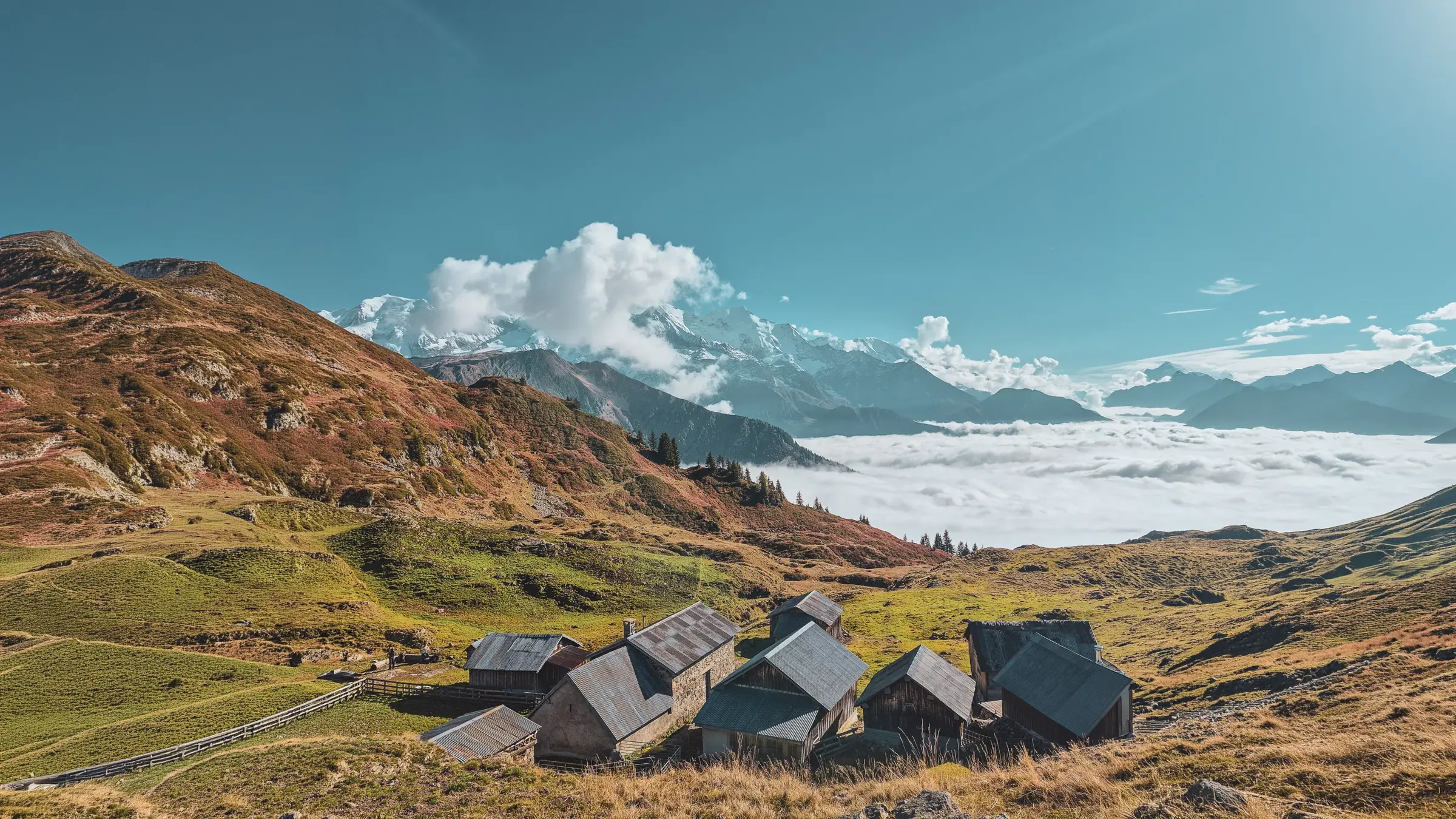 Wooden chalets overlook lush green valleys, facing snow-capped peaks.