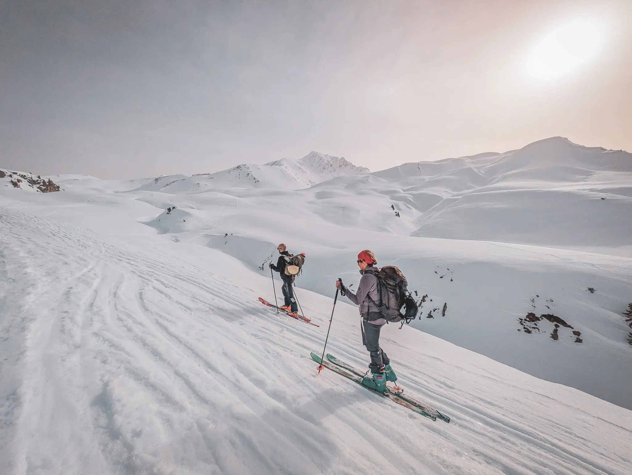 Two cross-country skiers advance across a snow-covered landscape, with majestic mountains in the background, under a clear sky.