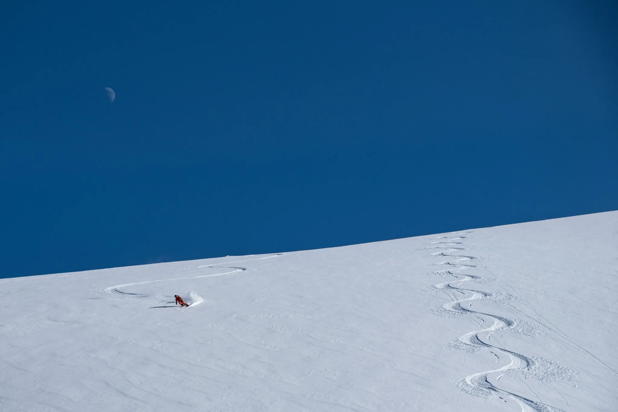 Un skieur descend une pente enneigée, laissant des traces derrière lui dans la neige immaculée. Le ciel est d'un bleu éclatant, et un croissant de lune est visible dans la partie supérieure gauche de l'image. Les