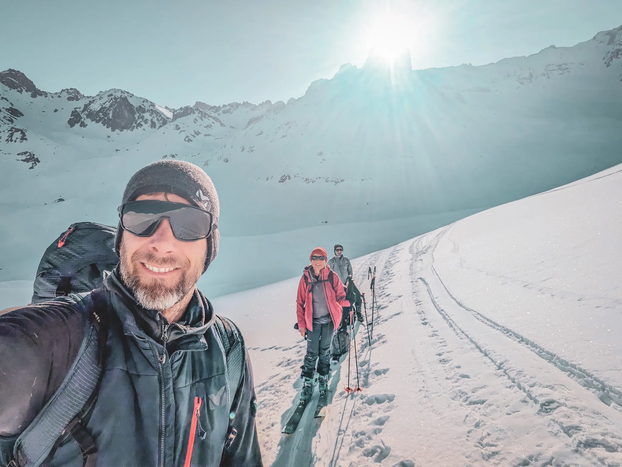 Three skiers on a snow-covered Alpine landscape, with the sun shining brightly in the background.