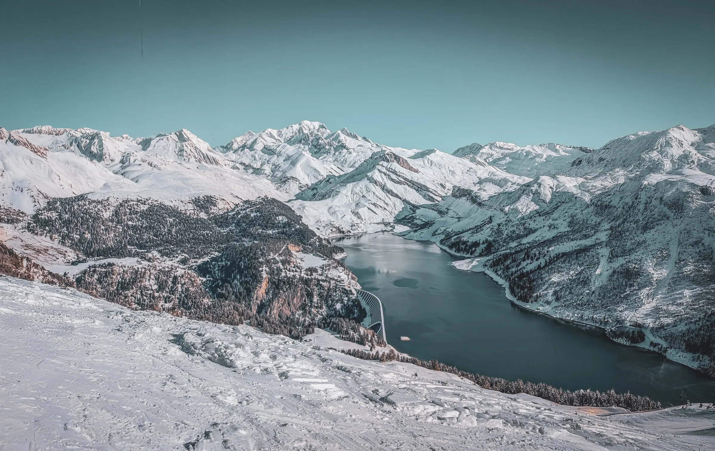 A snow-covered mountain panorama, with majestic peaks under a clear sky. In the centre, a calm lake reflects the surrounding mountains, while coniferous forests line the horizon.