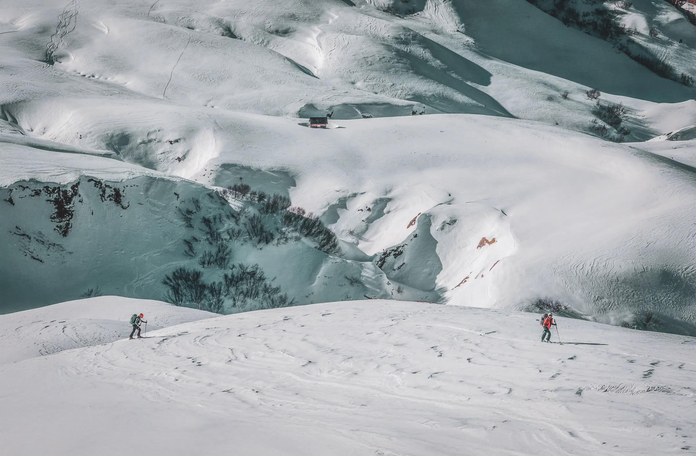 A snowy landscape with rolling hills covered in snow. Two skiers, one in a red jacket and the other in a dark jacket, are making their way down a slope. In the distance, a small cab