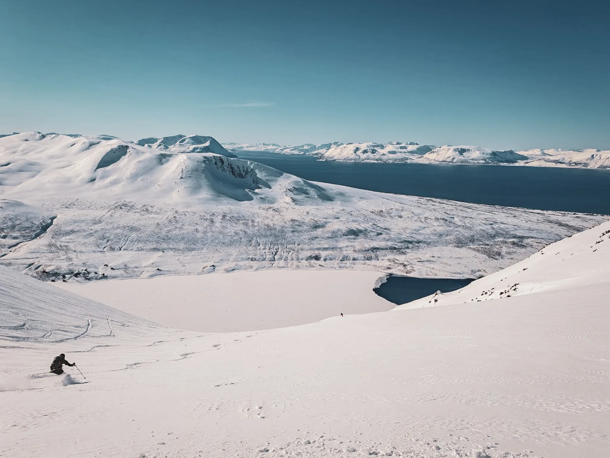 Des montagnes recouvertes de neige s'étendent à perte de vue, offrant un paysage hivernal majestueux. Un skieur descend une pente enneigée, laissant des traces derrière lui. Au loin, des sommets escarpés se