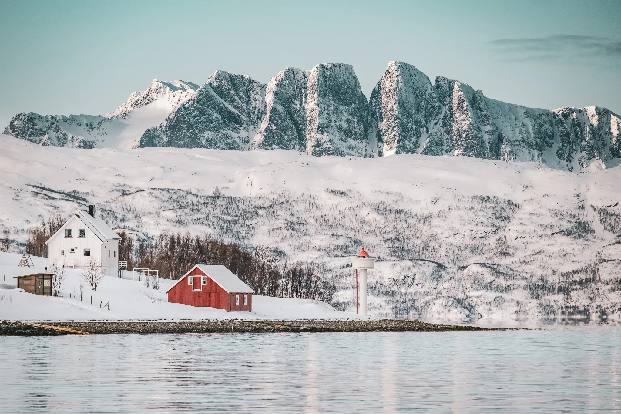 Un paysage hivernal avec des montagnes enneigées en arrière-plan, surplombant une étendue d'eau calme et réfléchissante. À gauche, une maison blanche est entourée de neige, tandis qu’à droite se trouve un petit bâtiment rouge