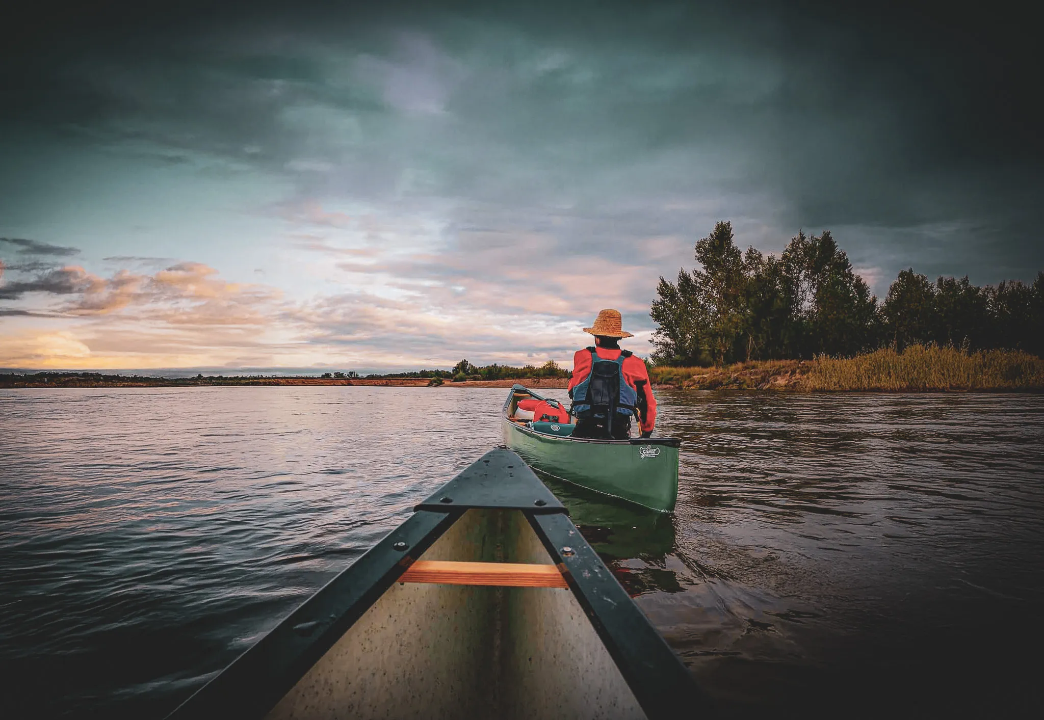 Un paysage aquatique paisible, avec une personne dans un canoë, dos à la caméra, paddle à la main. Cette personne porte un chapeau de paille, une veste rouge et un gilet de sauvetage. L’ar