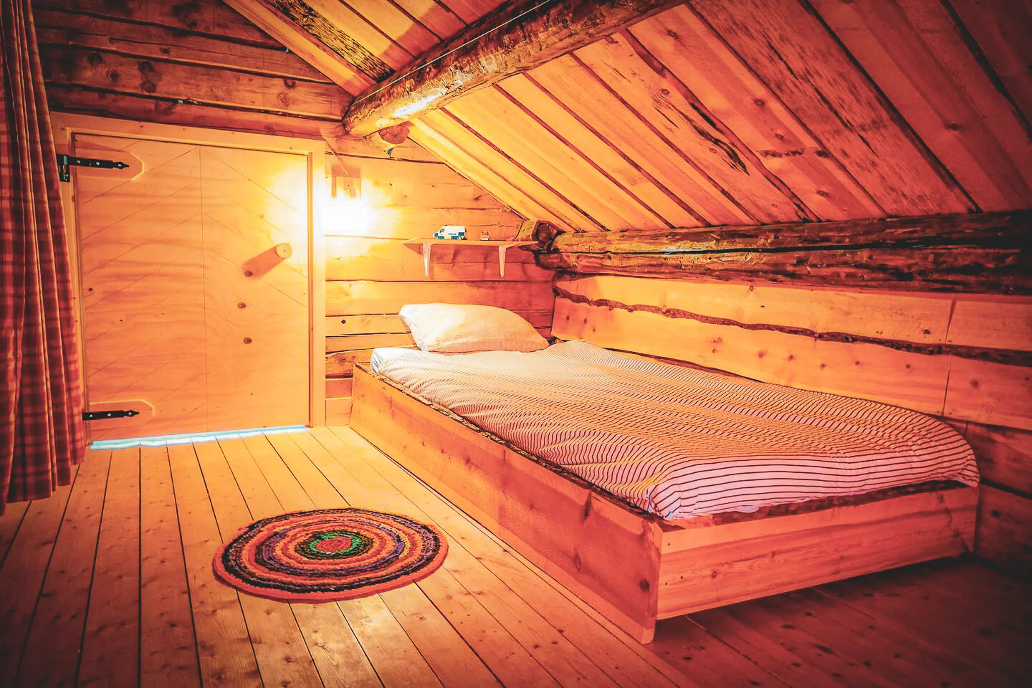 Une chambre en bois chaleureux avec des murs en planches de bois naturelles et un plafond incliné. Un lit en bois est placé contre le mur, recouvert d'une couette à rayures blanches et grises. Au sol, un