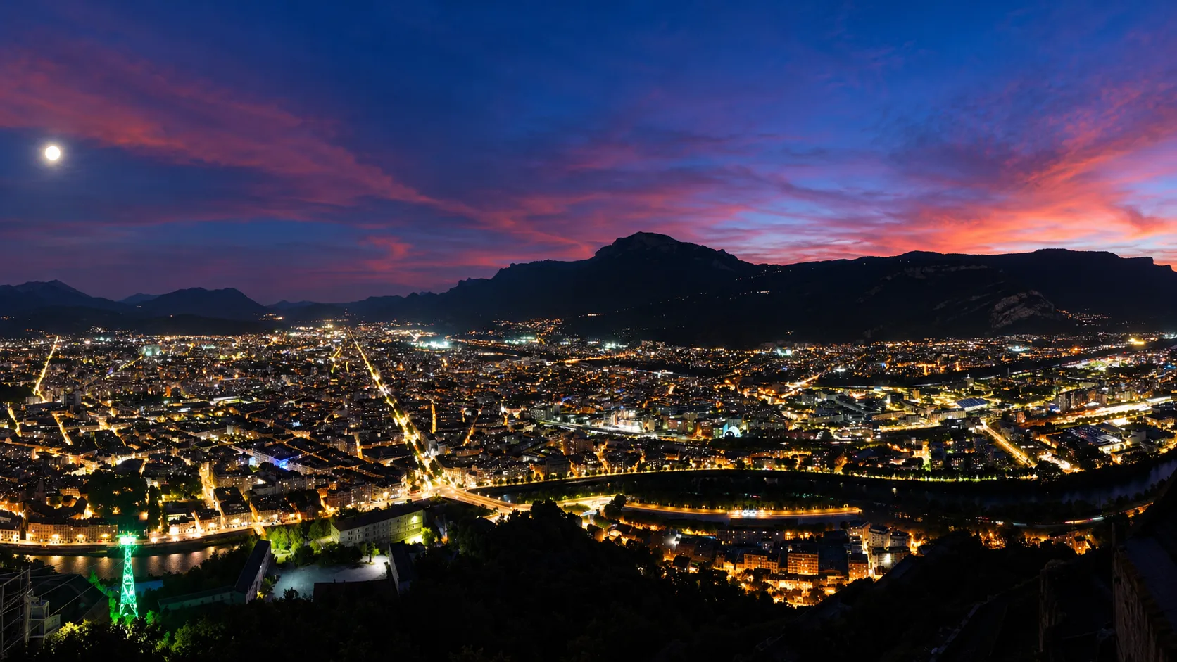 Vue panoramique de Grenoble illuminée la nuit, avec des montagnes en arrière-plan et un ciel coloré.