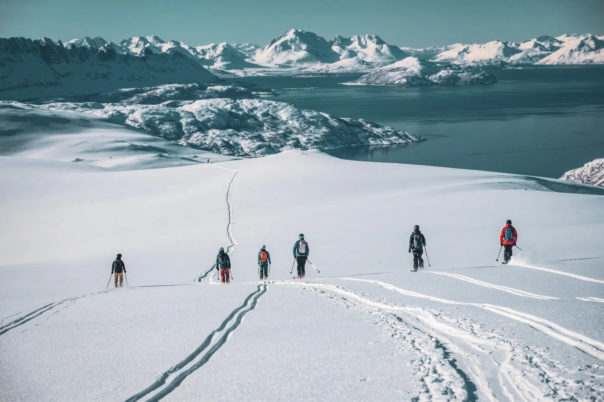 Un paysage enneigé avec un groupe de six alpinistes marchant sur une pente. La neige immaculée est parcourue de traces laissées par leurs skis, menant vers un horizon montagneux impressionnant. En arrière-plan, des