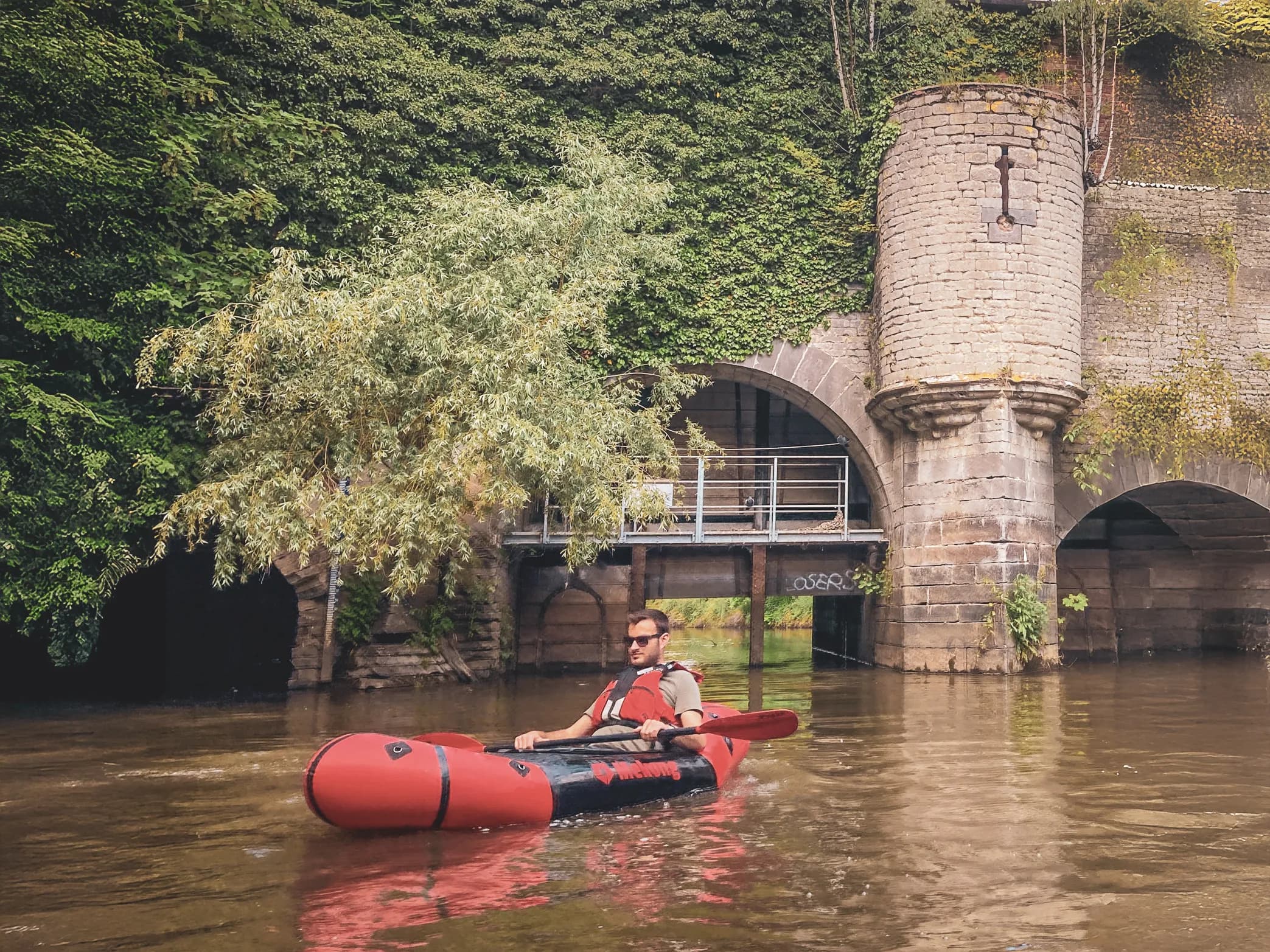 A packraft paddler cruises peacefully under an ancient, green bridge in Belgium.
