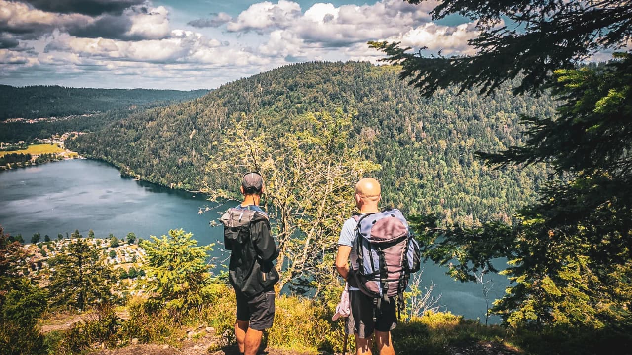 Two hikers admire a green panorama and a serene lake in the sunny Vosges mountains.