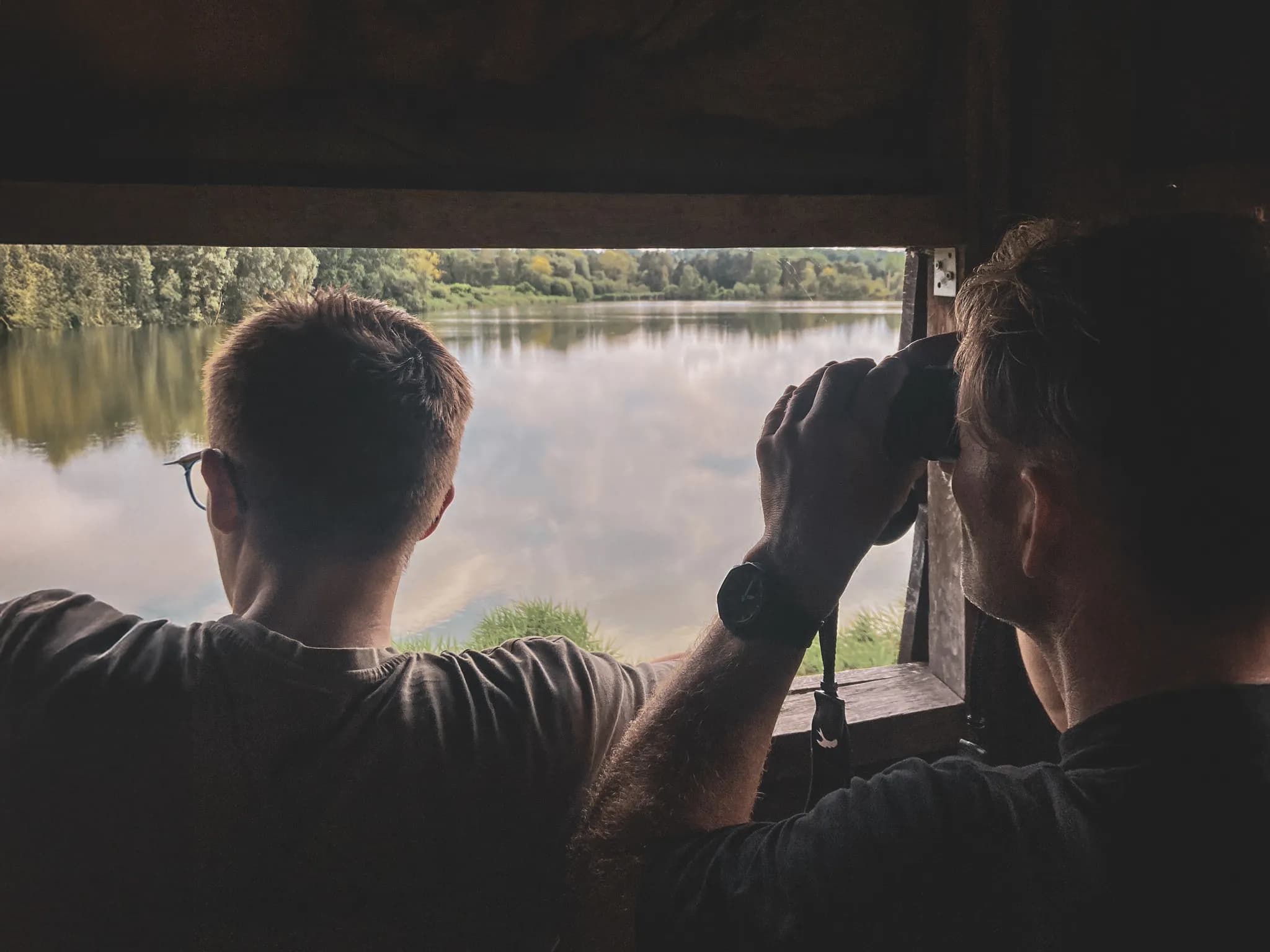 Two people peacefully observing a green lake through a window, equipped with binoculars.
