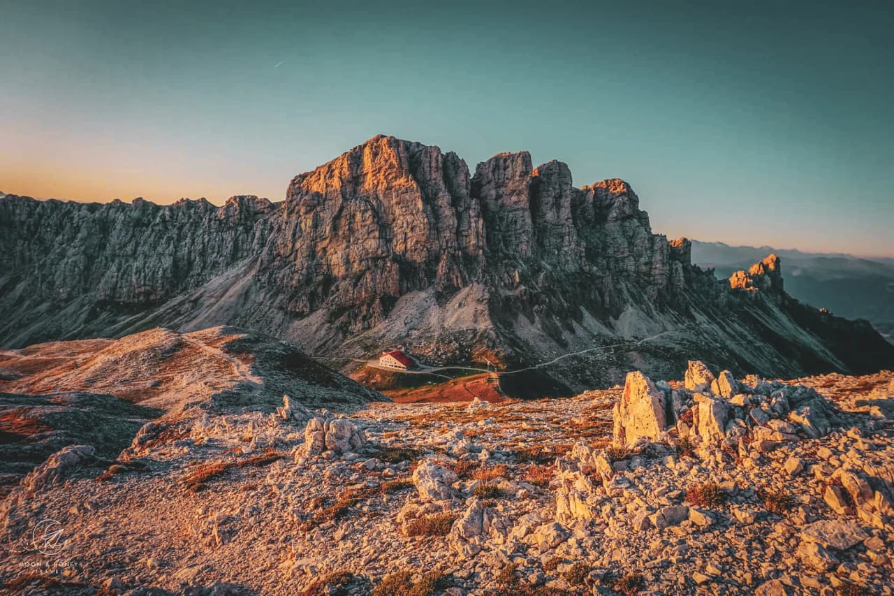 An impressive mountain landscape is visible, with sheer, rocky cliffs rising majestically beneath a pastel-hued sky. The sun sets, lighting up the peaks in a golden glow. Visit