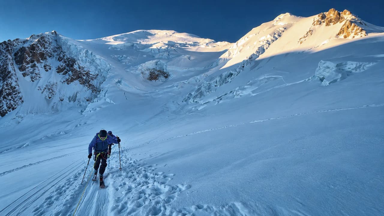 Un skieur grimpe sur une pente enneigée, avec le majestueux Mont Blanc en arrière-plan.