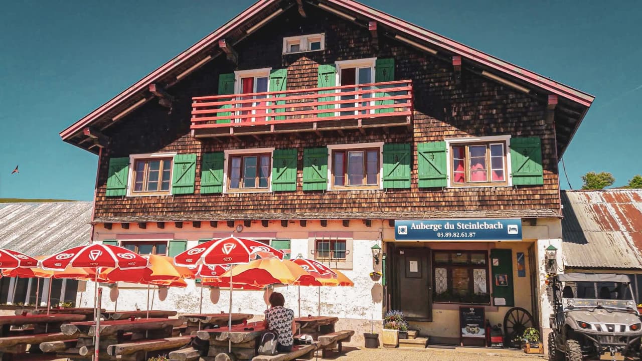 Auberge charmante avec balcon, parasols rouges, idéale pour une pause en randonnée dans les Vosges.