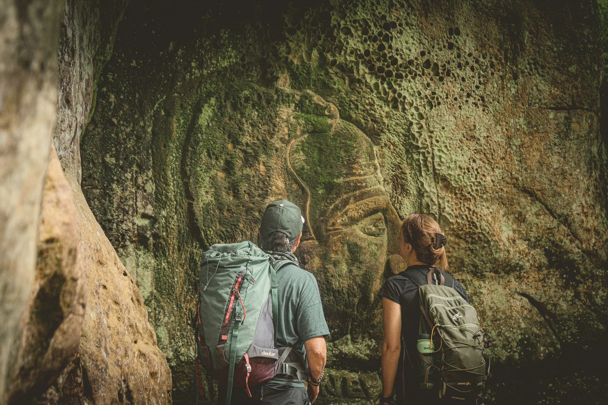 Deux randonneurs admirent des sculptures anciennes sur une paroi rocheuse verdoyante en Petite Suisse luxembourgeoise.