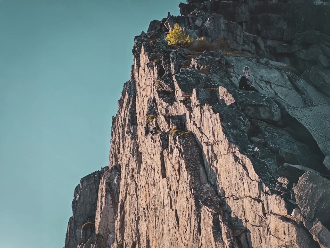 Een persoon die op een steile klif zit, omringd door een prachtig landschap en een strakblauwe lucht.