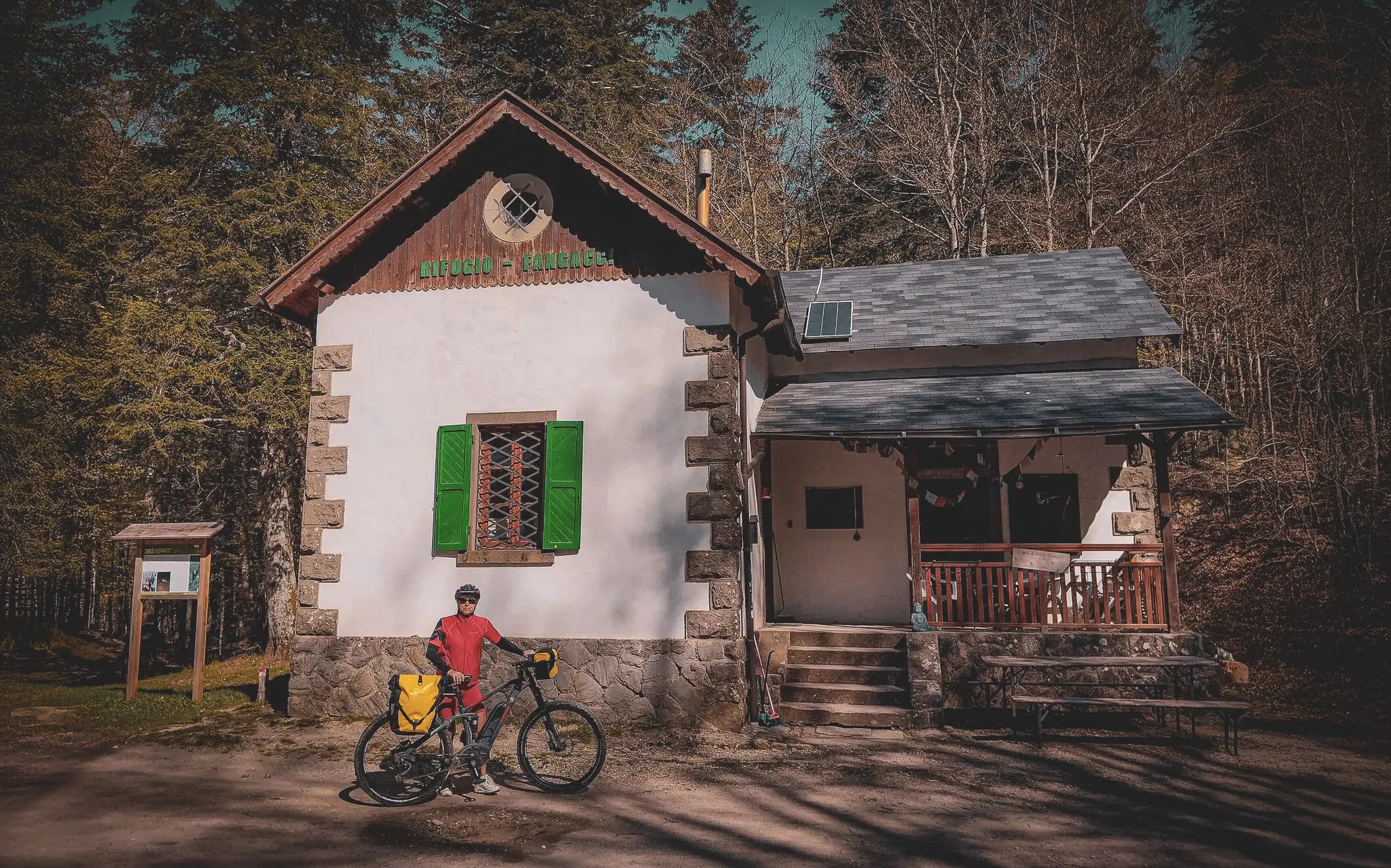 Un chalet en pleine nature, construit en pierre et en bois, avec des fenêtres à volets verts. À l'avant, une personne en tenue de cyclisme se tient à côté d'un vélo chargé.