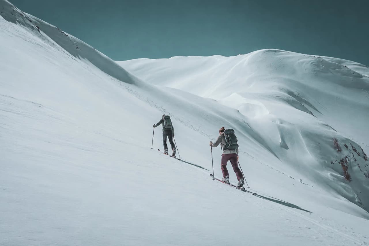 Two ski tourers are making their way through majestic snow-covered landscapes beneath a blue sky.